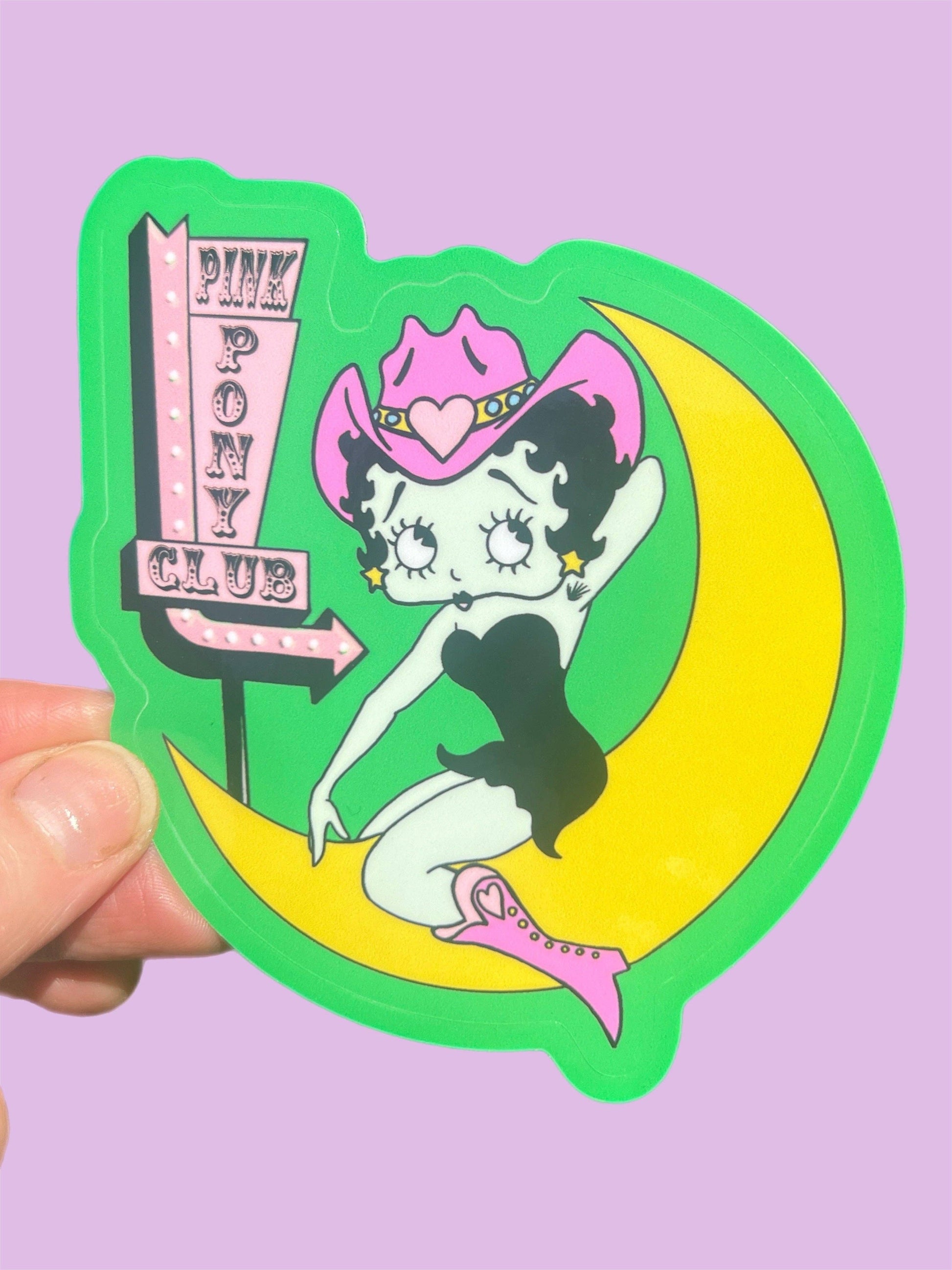 Sticker of betty boop on a moon with 'Pink Pony Club' sign, held against a purple background.