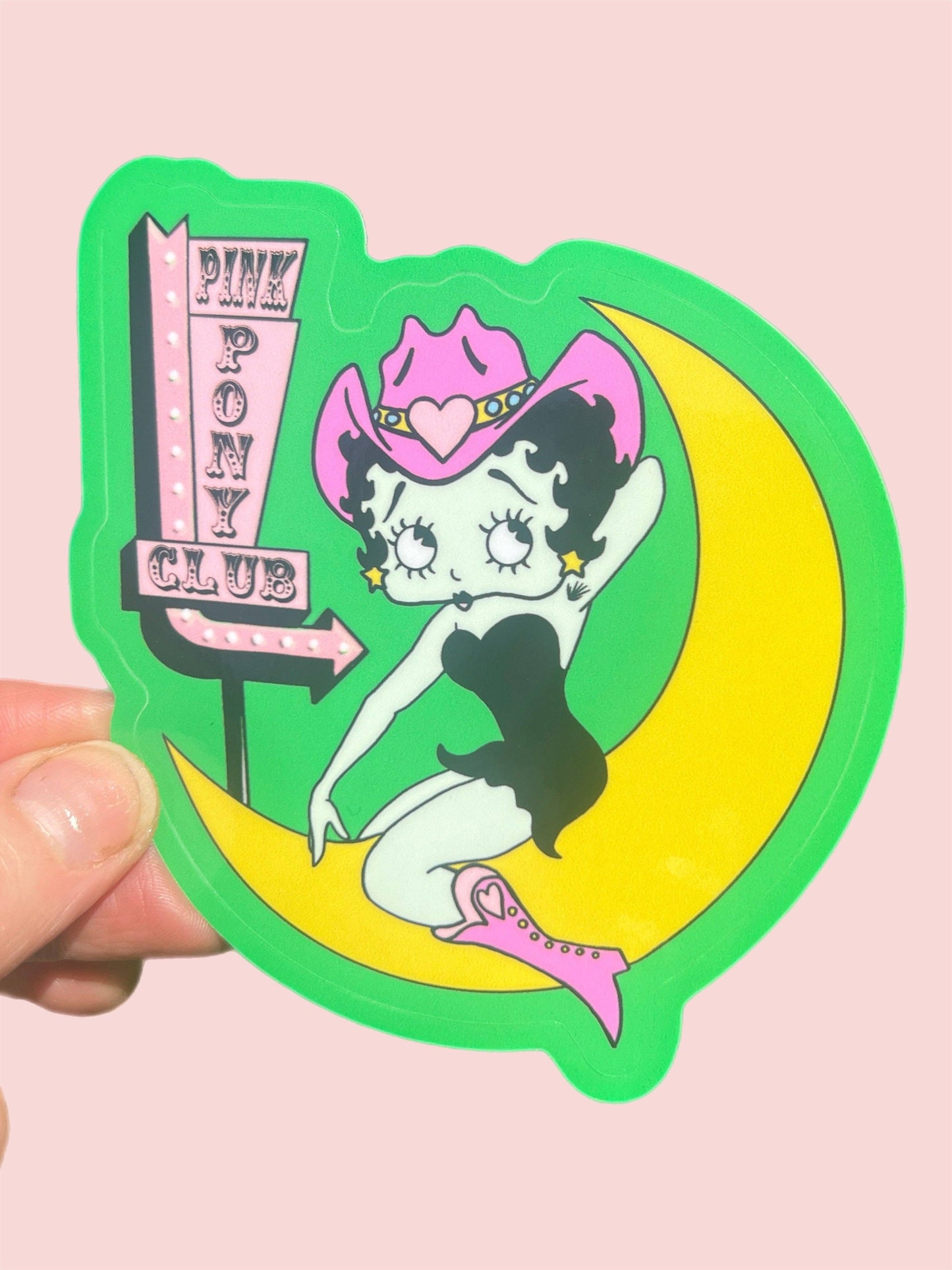 Sticker of a betty boop on a moon with 'Pink Pony Club' sign, held against a pink background.