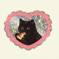 Sticker of a black cat holding a pizza inside a heart-shaped frame with a pink border on a beige background.