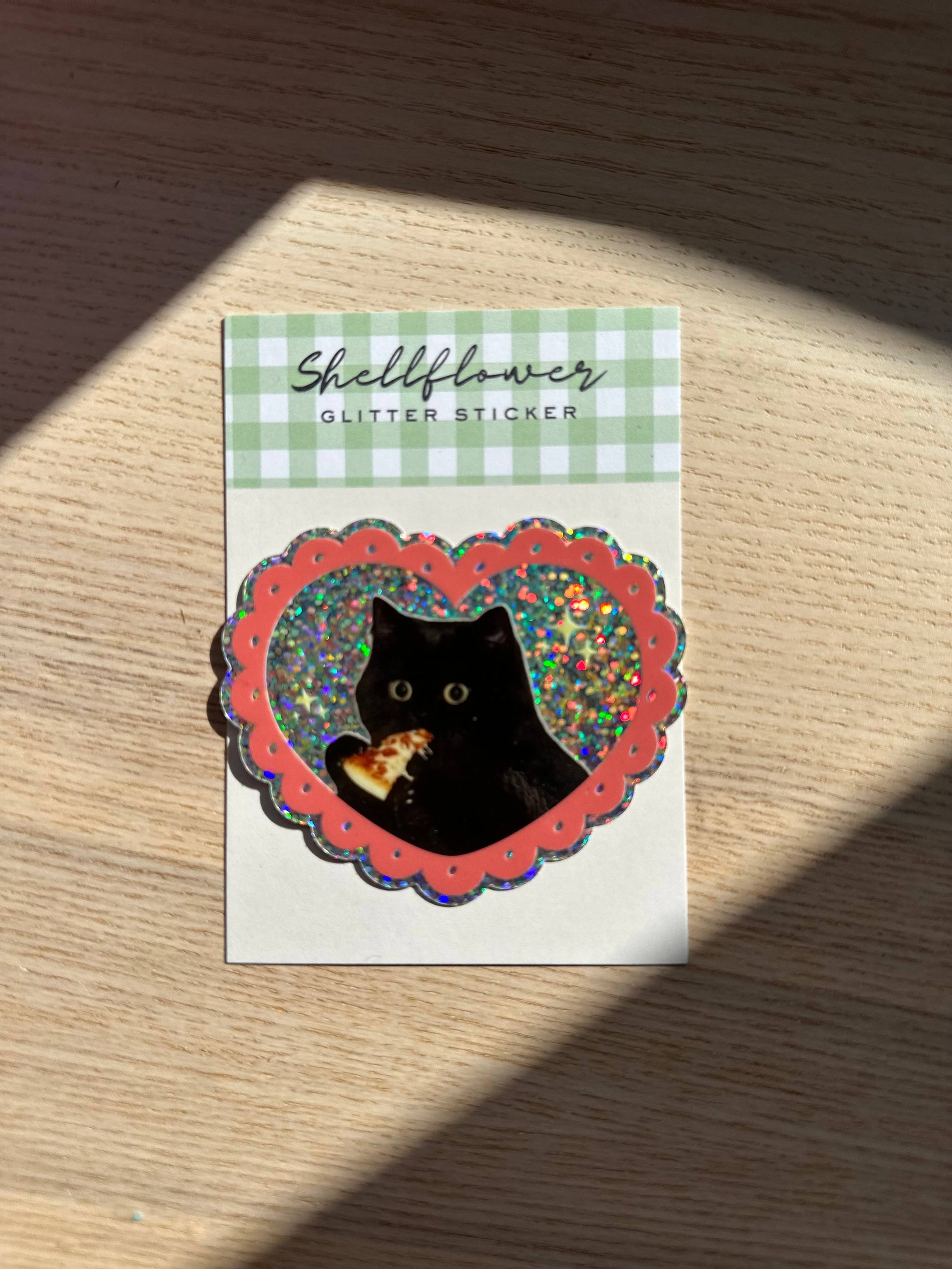 Sticker with a black cat eating pizza on a checkered background, brand 'Shellflower' visible