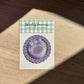 Purple glitter sticker on a card with 'Shellflower' branding on a wooden surface