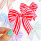 Red and pink striped bow held by a hand with teal nail polish against a colorful background.