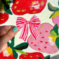 Hand holding a pink bow over a colorful fabric background with strawberries and flowers.