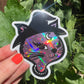 Holographic cat sticker with witch hat held in front of green leaves