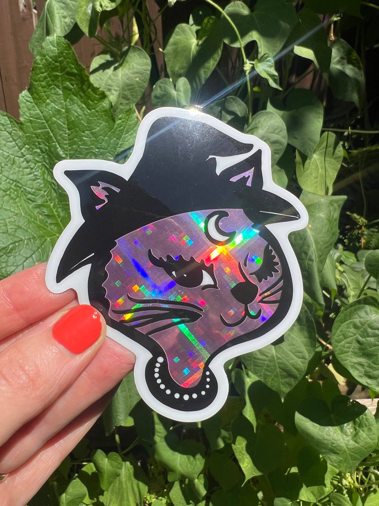 Holographic cat sticker with witch hat held in front of green leaves
