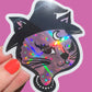 Holographic cat sticker with witch hat held by a hand on a pink background