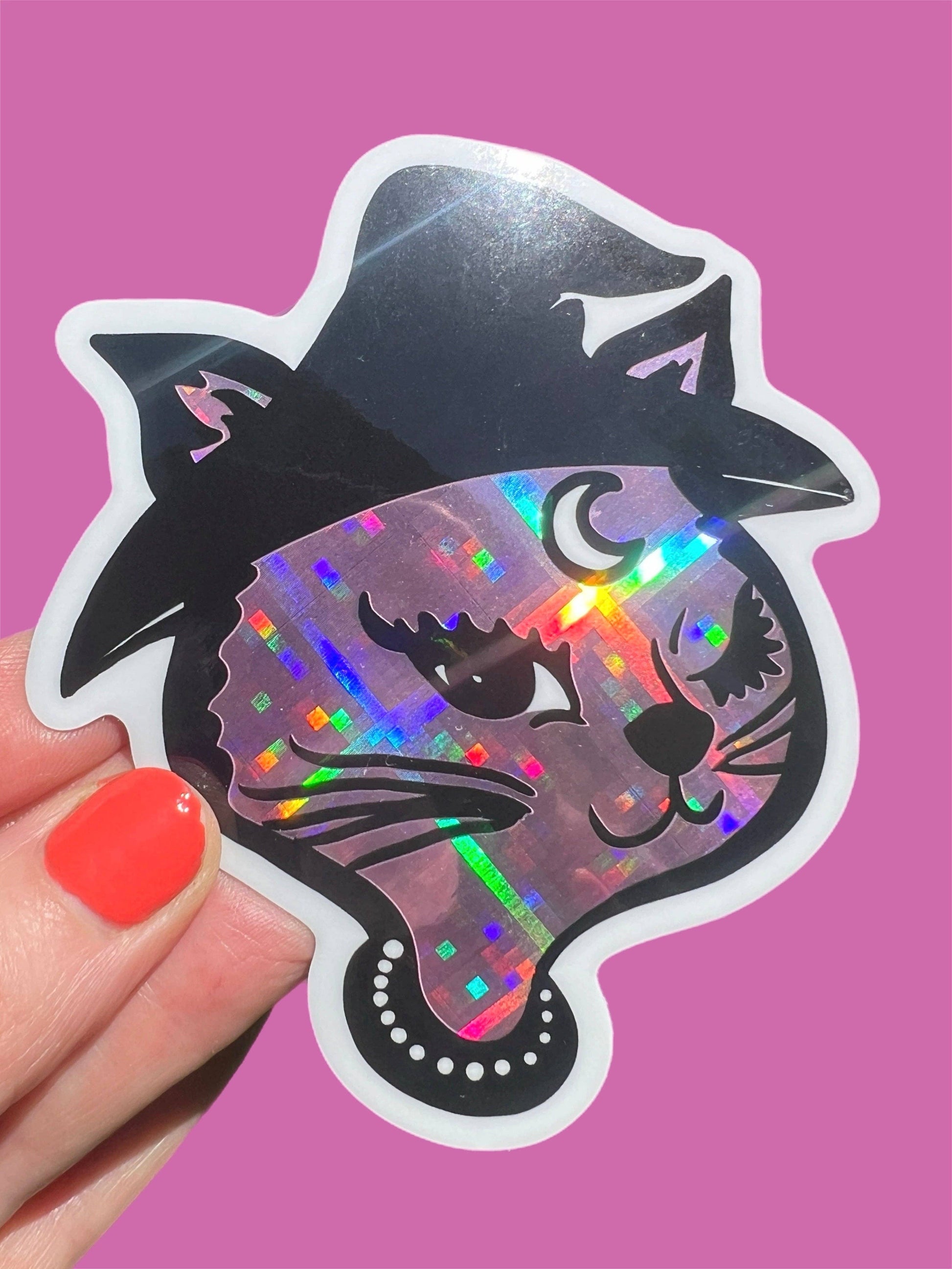 Holographic cat sticker with witch hat held by a hand on a pink background