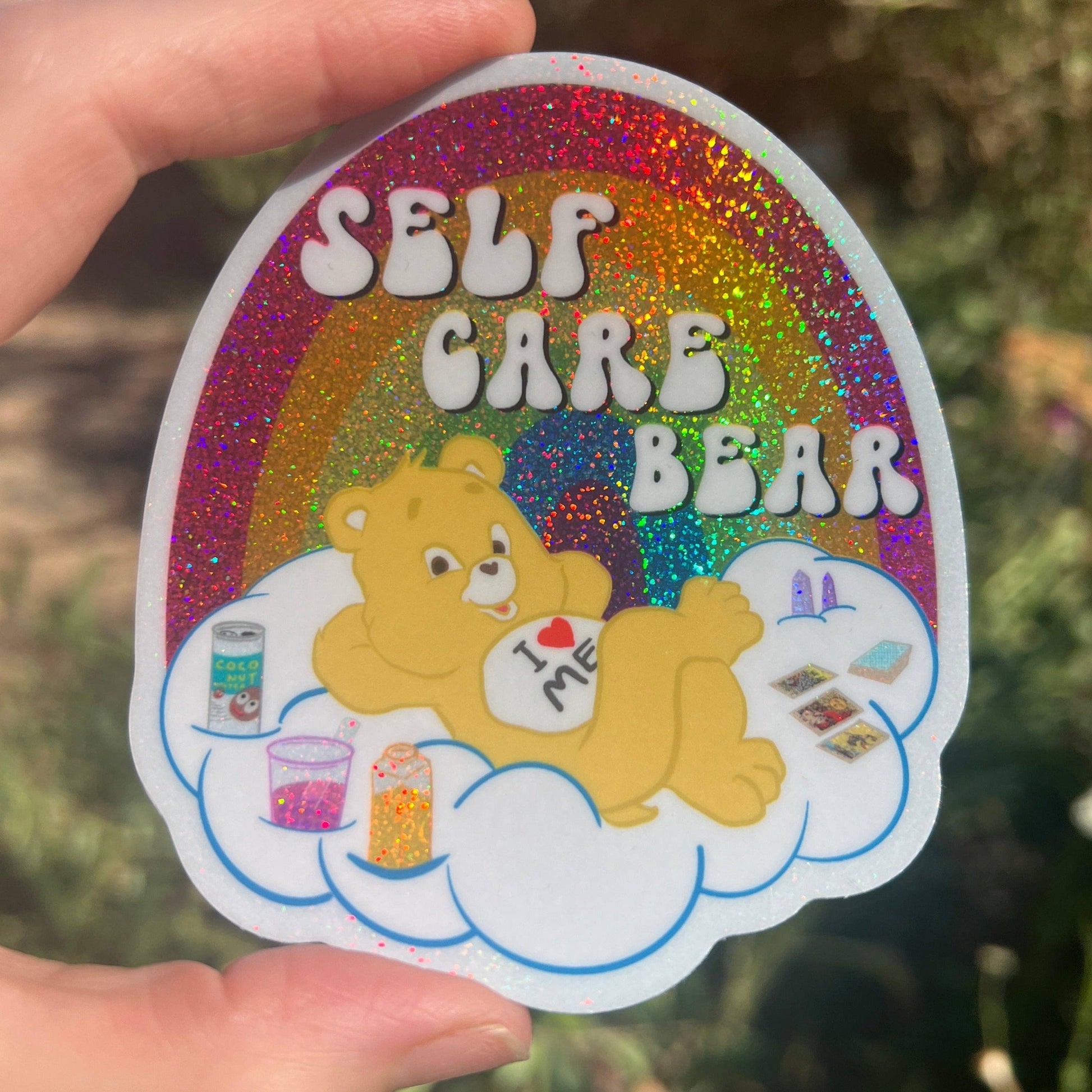 Sticker with 'Self Care Bear' design held by a hand against a blurred natural background