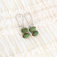 A pair of silver drop earrings with two green glass olives, displayed on a cream-colored ruffled fabric background.