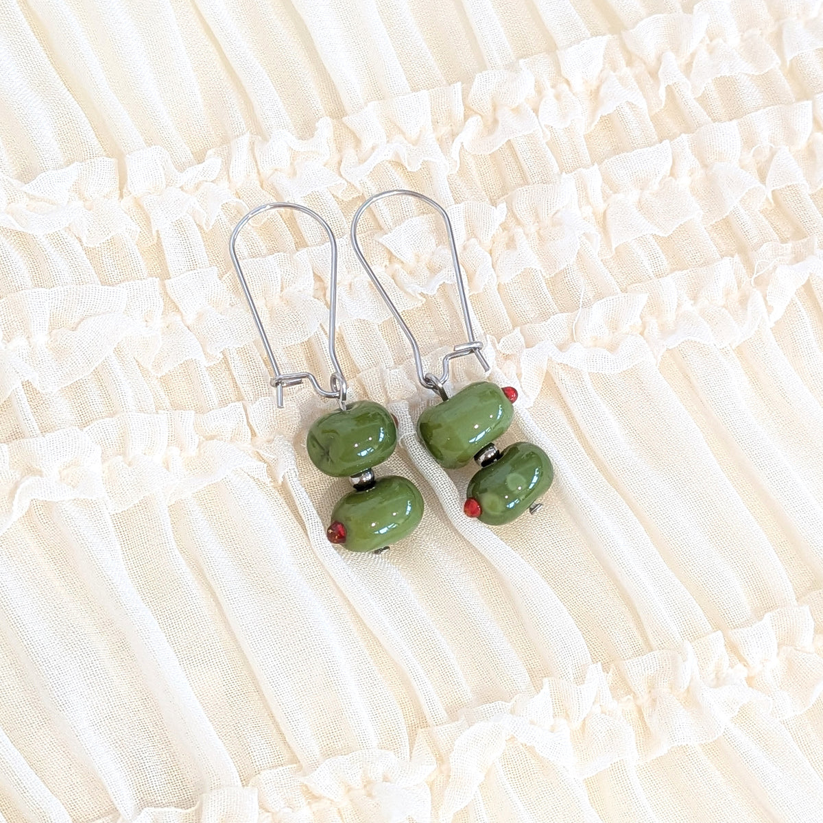A pair of silver drop earrings with two green glass olives, displayed on a cream-colored ruffled fabric background.
