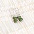 A pair of silver drop earrings with two green glass olives, displayed on a cream-colored ruffled fabric background.