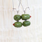 A pair of handmade dangle earrings with glossy olive green glass beads hanging from silver wire hooks against a soft, textured off-white background.
