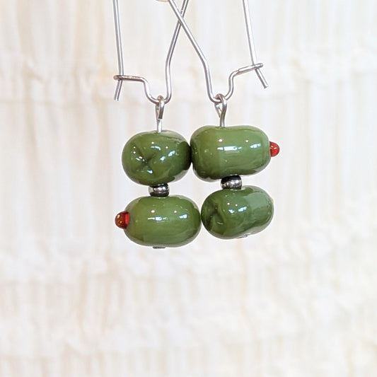 A pair of handmade dangle earrings with glossy olive green glass beads hanging from silver wire hooks against a soft, textured off-white background.