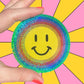 Hand holding a colorful smiley face sticker against a multicolored background