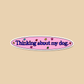 An oval sticker with the text 'Thinking about my dog.' in dark blue script, set on a pink background with small multicolored star accents and a white border, displayed on a solid beige background.