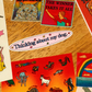 A collection of colorful stickers displayed on a wooden tabletop. The stickers feature various designs including a glittery cat, a red graphic with the text 'THE WINNER TAKES IT ALL', a pink oval sticker reading 'Thinking about my dog.