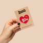 Hand holding a 'Scout's Honour' card with a heart-shaped badge on a beige background