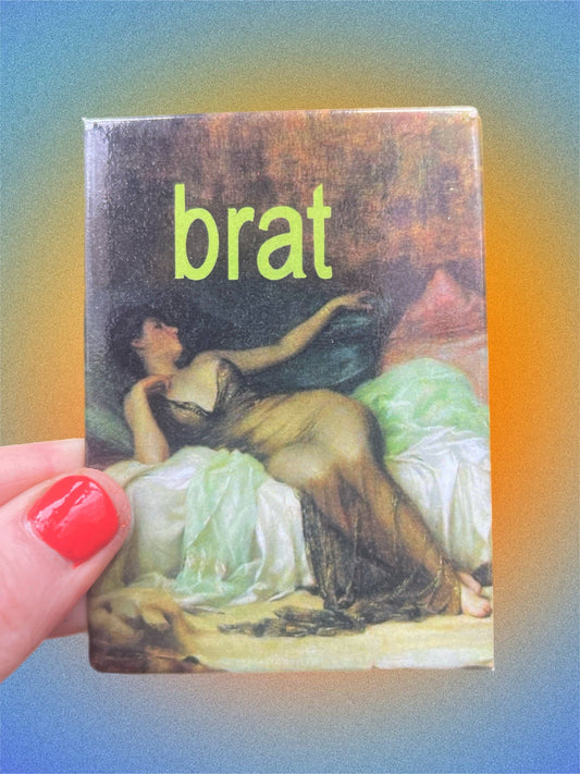 magnet with a classical painting cover and the word 'brat' in green text, held by a hand with red nail polish.