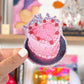 Hand holding a decorative sticker with a pink cake design and cherries against a colorful, blurred background.