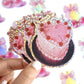 Sticker of a pink cake with cherries held by a hand against a colorful, blurred background.