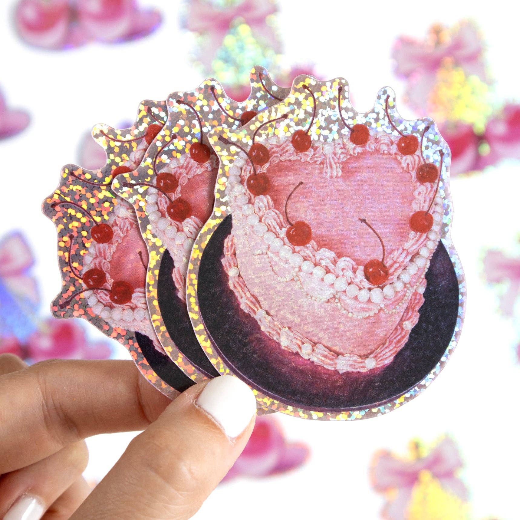 Sticker of a pink cake with cherries held by a hand against a colorful, blurred background.