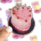 Heart-shaped cake with cherries and a crown on a reflective surface, held by a hand.