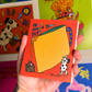 A hand holding a small illustrated notebook or card with a bright orange cover, featuring a large yellow cheese wedge and the text 'IS IT COFFEE TIME YET?' against a colorful, cartoon-filled background.