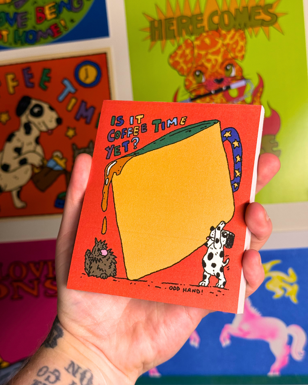 A hand holding a small illustrated notebook or card with a bright orange cover, featuring a large yellow cheese wedge and the text 'IS IT COFFEE TIME YET?' against a colorful, cartoon-filled background.