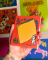 A hand holding a small illustrated notebook or card with a bright orange cover, featuring a large yellow cheese wedge and the text &
