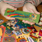 A person holds a green rectangular sticker with retro computer graphics and the text 'Move! It's Computer Time!' over a pile of colorful vintage-style stickers on a wooden surface.