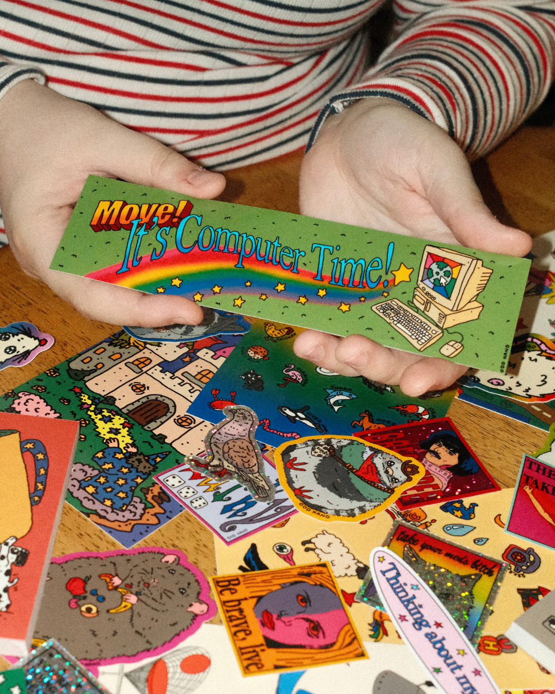 A person holds a green rectangular sticker with retro computer graphics and the text 'Move! It's Computer Time!' over a pile of colorful vintage-style stickers on a wooden surface.