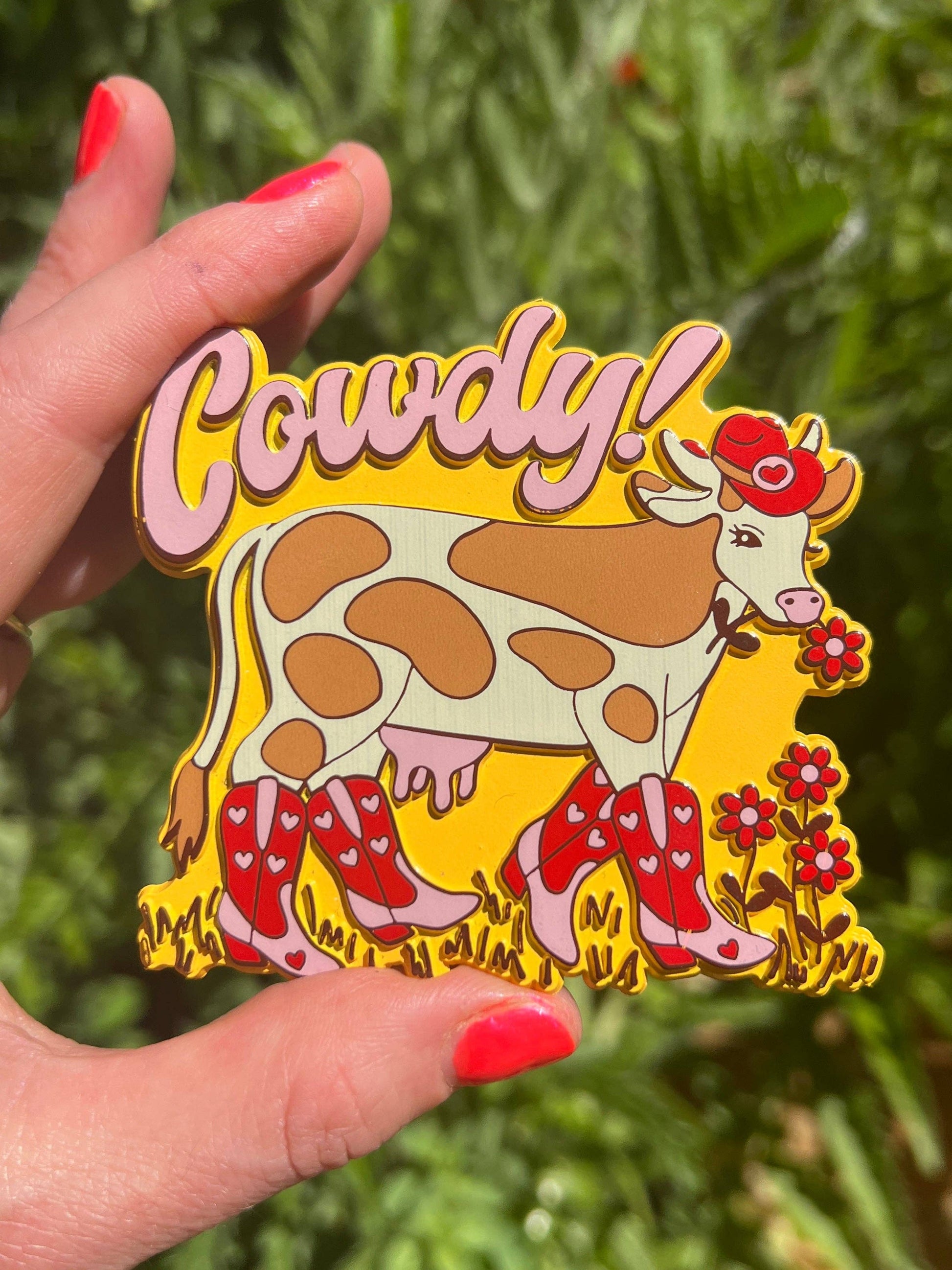 Cowdy! magnet held by a hand with red nail polish against a green leafy background