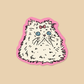A die-cut sticker featuring a fluffy white cat with blue eyes and a pink bow, outlined in black and set against a pink border, displayed on a solid beige background.