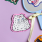 A styled flat-lay featuring a central die-cut sticker of a fluffy white cat with blue eyes and a pink bow on a pink border, set against a lavender background. A light blue hand mirror with a cream ribbon is positioned to the right, alongside partial views of other colorful cat stickers.