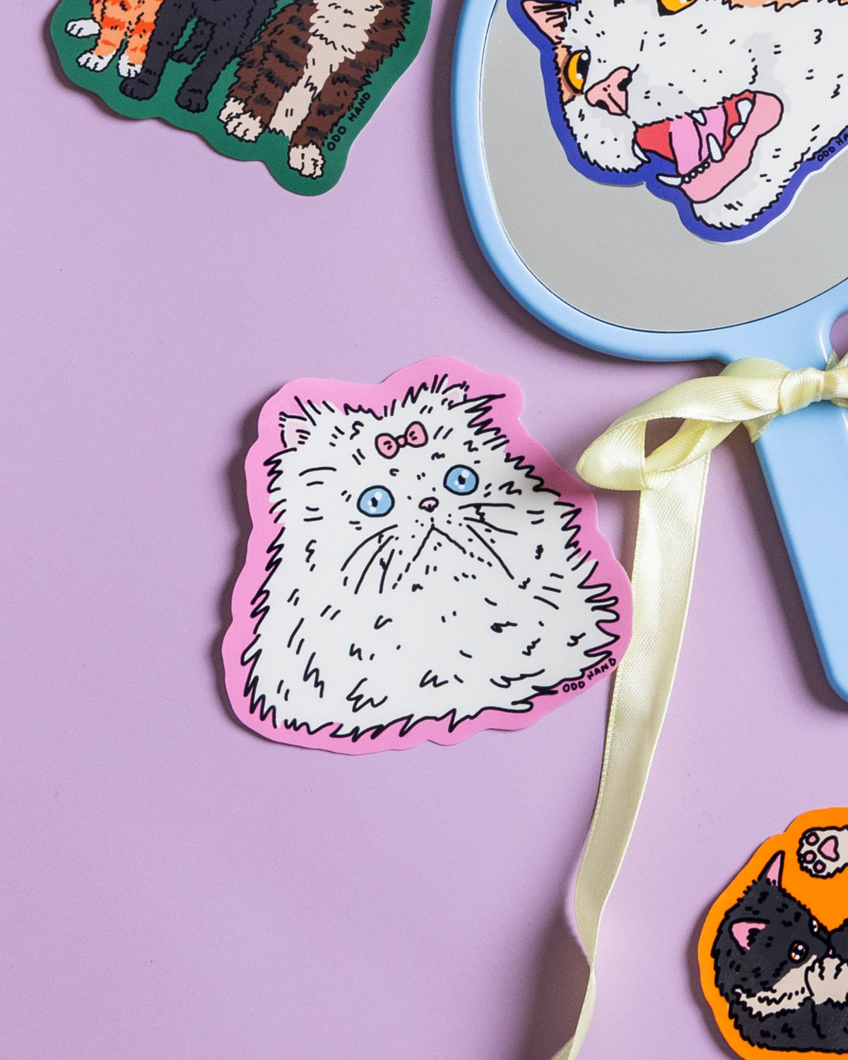 A styled flat-lay featuring a central die-cut sticker of a fluffy white cat with blue eyes and a pink bow on a pink border, set against a lavender background. A light blue hand mirror with a cream ribbon is positioned to the right, alongside partial views of other colorful cat stickers.