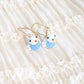 A pair of gold-tone dangle earrings with small white bird charms displayed on a cream-colored ruffled fabric background.