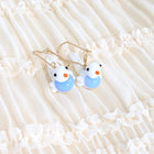 A pair of gold-tone dangle earrings with small white bird charms displayed on a cream-colored ruffled fabric background.