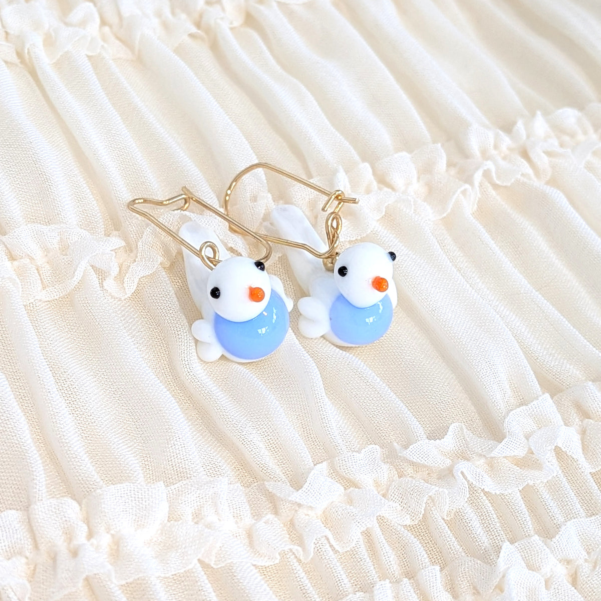 A pair of gold-tone dangle earrings with small white bird charms displayed on a cream-colored ruffled fabric background.