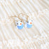 A pair of gold-tone dangle earrings with small white bird charms displayed on a cream-colored ruffled fabric background.