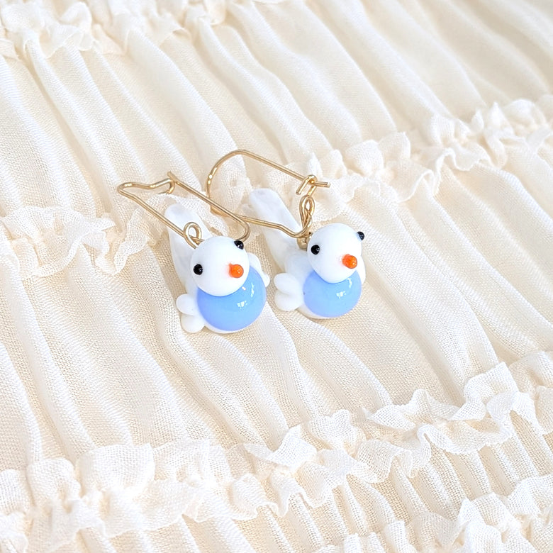 A pair of gold-tone dangle earrings with small white bird charms displayed on a cream-colored ruffled fabric background.