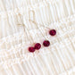 Gold-tone drop earrings with glossy deep red bead drops, displayed on a cream-colored ruffled fabric background.