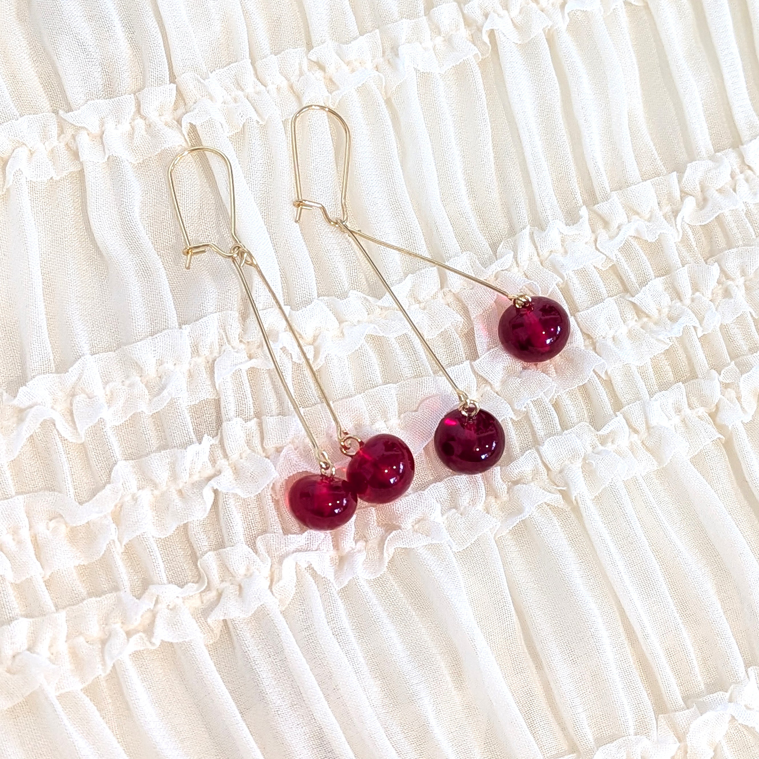 Gold-tone drop earrings with glossy deep red bead drops, displayed on a cream-colored ruffled fabric background.