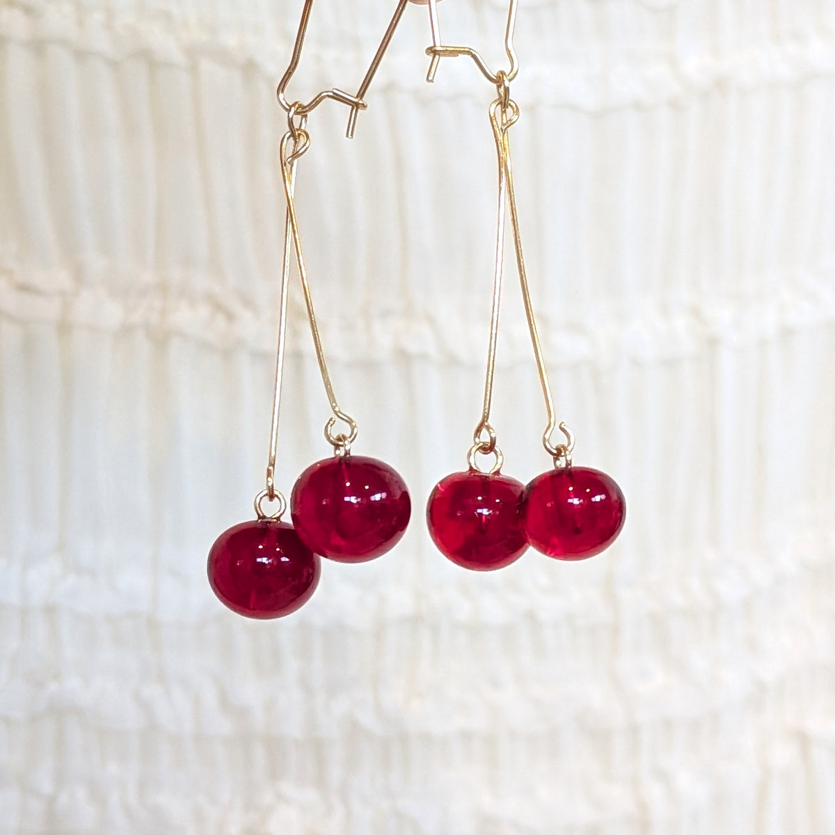 Gold-tone dangle earrings with glossy deep red bead drops each, hanging against a soft white textured fabric background.
