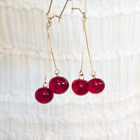 Gold-tone dangle earrings with glossy deep red bead drops each, hanging against a soft white textured fabric background.