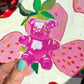 Pink glitter gummy bear sticker held by a hand against a floral background.