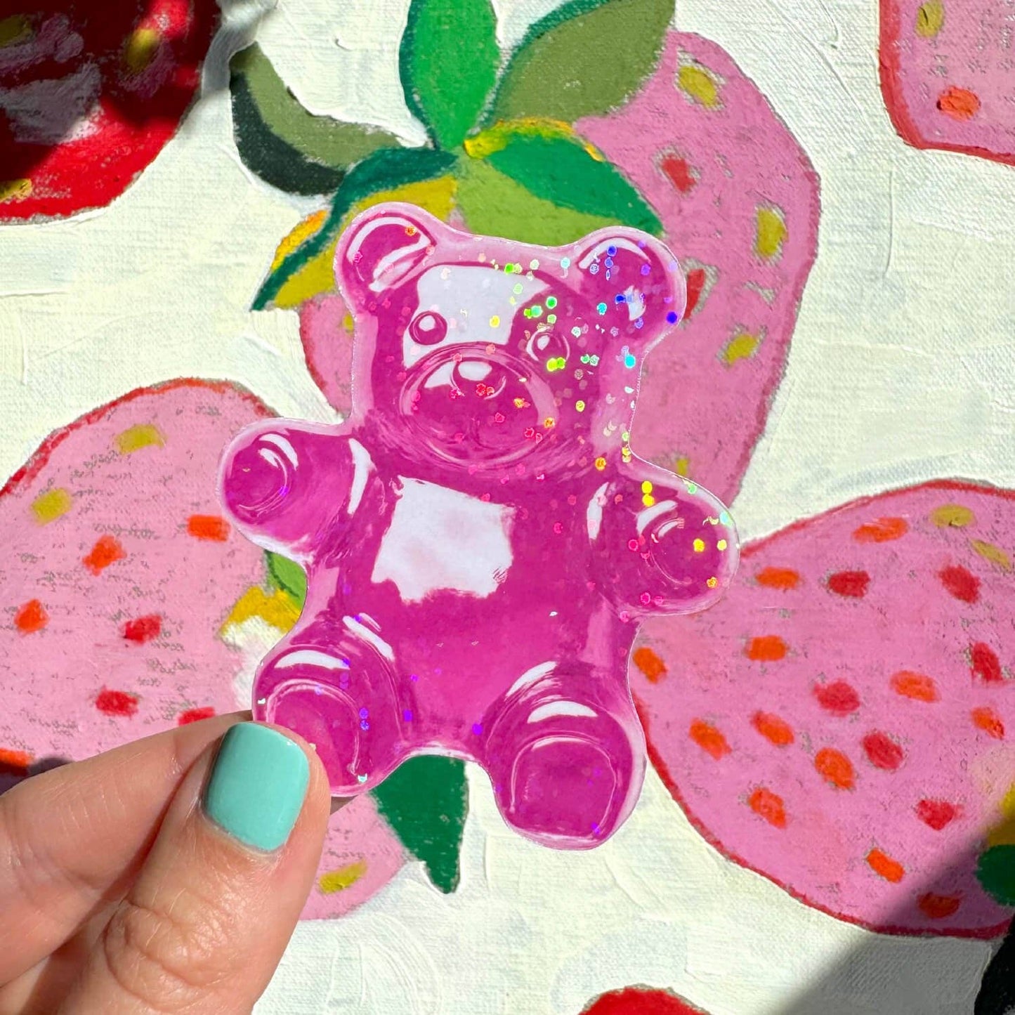 Pink glitter gummy bear sticker held by a hand against a floral background.