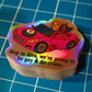 A stack of die-cut stickers featuring a pink convertible car with two women and the text 'Hop in diva, we're going to be gay & do crime.' The stickers have an iridescent holographic finish and are photographed on a blue grid cutting mat.