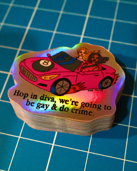 A stack of die-cut stickers featuring a pink convertible car with two women and the text 'Hop in diva, we're going to be gay & do crime.' The stickers have an iridescent holographic finish and are photographed on a blue grid cutting mat.