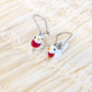 A pair of silver fishhook earrings with small white bird charms featuring red bellies, orange beaks, and black eyes, displayed on a cream-colored ruffled fabric background.