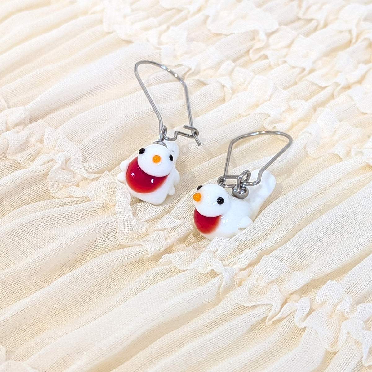 A pair of silver fishhook earrings with small white bird charms featuring red bellies, orange beaks, and black eyes, displayed on a cream-colored ruffled fabric background.